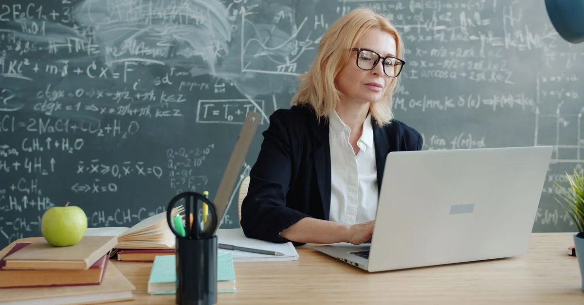 Un professeur travaille sur un ordinateur portable dans une salle de classe où des équations sont inscrites au tableau noir, concentré et professionnel.