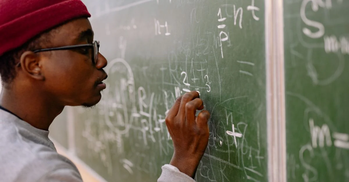 Un jeune homme portant des lunettes et un bonnet rouge résolvant des problèmes de mathématiques sur un tableau noir.