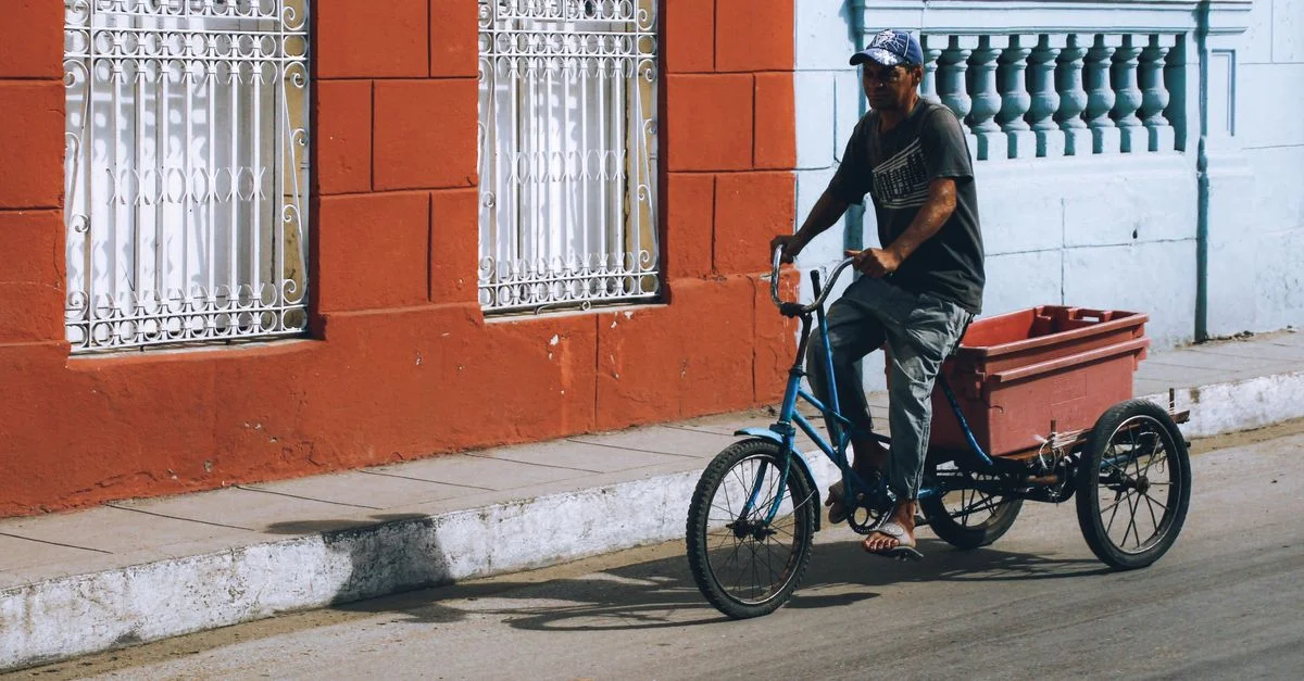 Un homme roule sur un tricycle avec une remorque le long d'une rue urbaine vibrante aux façades colorées.