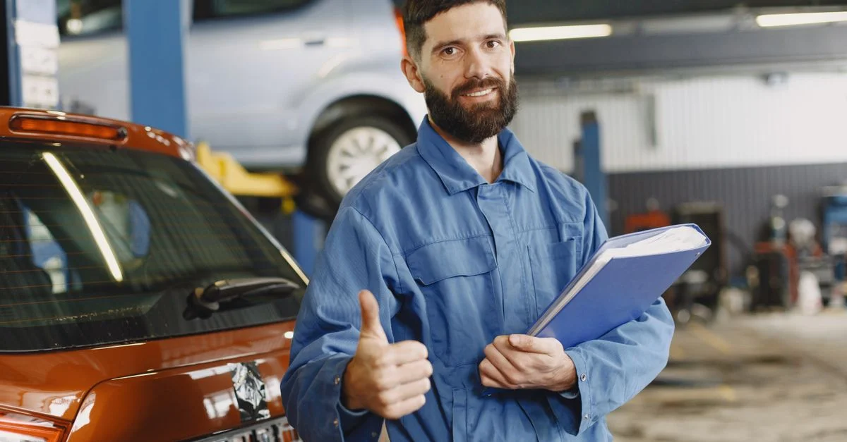 Mécanicien automobile en salopette bleue avec un clipboard faisant un signe de pouce levé dans le garage.